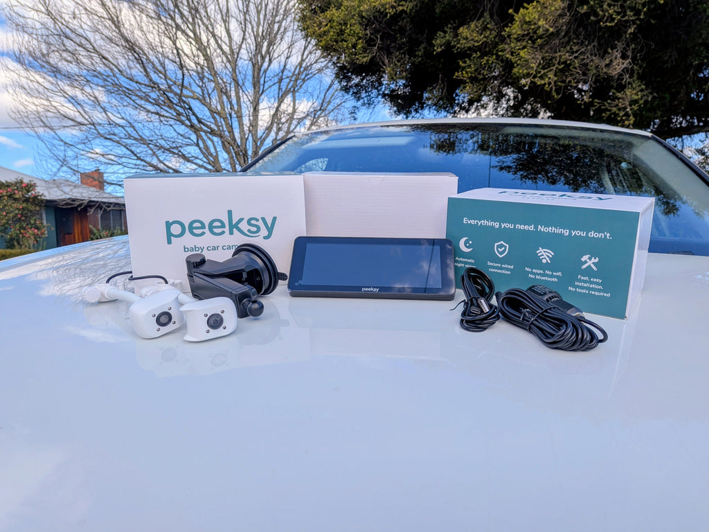Peeksy baby car camera kit laid out on the hood of a white car outdoors, including two white cameras, a black display screen, cables, and branded product boxes.