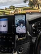 A Peeksy baby monitor tablet mounted on a car dashboard air vent, showing a baby in a car seat inside a vehicle cabin.