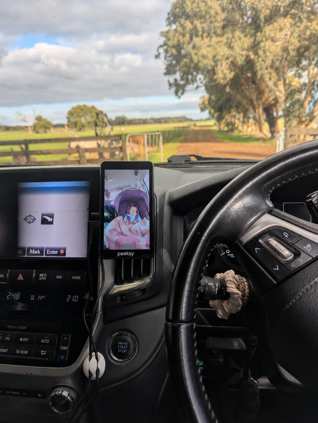Peeksy mini mounted to an air vent in the cars dash, showing a baby in a car seate, with the car's steering wheel and center console visible. A real outdoor landscape is seen through the windshield.