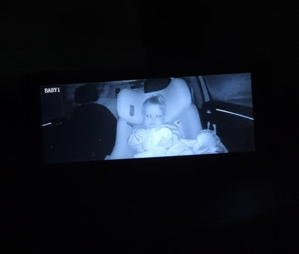 A dark screen showing a monochrome night-vision photo of a baby sitting in a car seat inside a vehicle, with the text 'BABY1' in the top-left corner of the screen.
