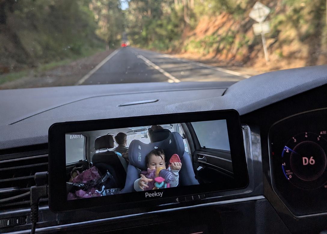 A car's infotainment screen displays a video feed of a child sitting in a car seat inside the vehicle, with a real road and trees visible through the windshield. The screen shows the brand name Peeksy at the bottom.
